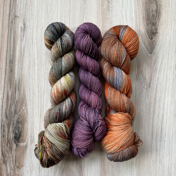 Three skeins of yarn in orange, gray and gold speckle, solid purple and variegated orange and gray. 